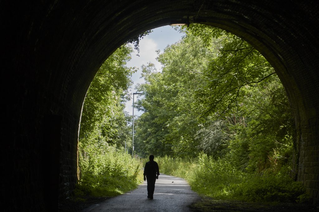 Oinezko bat tunel batetik irteten, tunelaren barnetik ikusita. Kanpoan Landare ugari dago. Pertsonaren silueta edo itzala baino ez da ikusten.