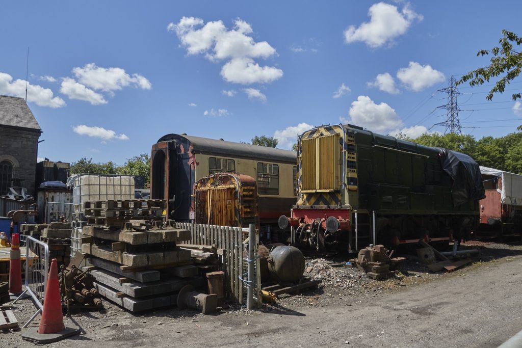 Bi tren-makina zahar eta txiki, eta trenbidearekin lotutako beste hamaika traste (hormigoizko trenbide-trabesak, hesiak, metalezko piezak…). Zeruan hodei gutxi batzuk daude.