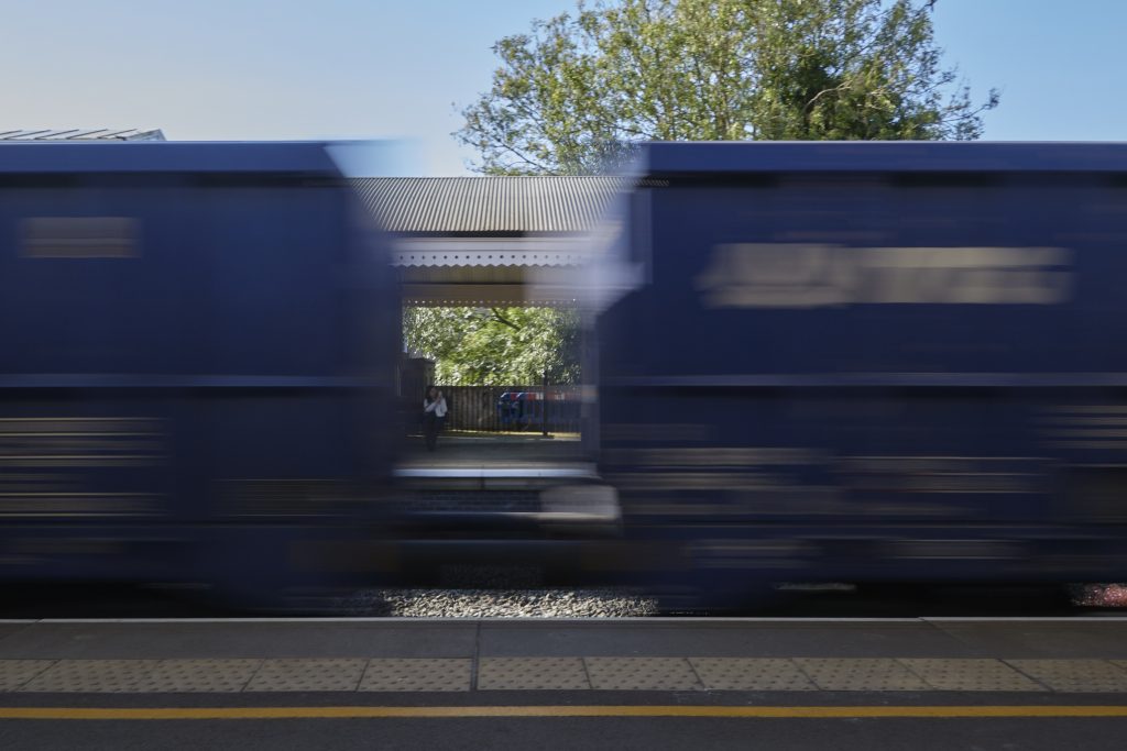 Pertsona bat tren-geltokiko pareko nasan. Pertsona tren-geltokitik pasatzen ari den merkantzia trenari argazki bat egiten ari zatzaio. Tren horrek daramatzan bi kontainer urdinen arteko tartetik egin zaio argazkia pertsonari. Pertsona eta nasa aratz ikusten dira, baina igarotzen ari den trena mugituta.